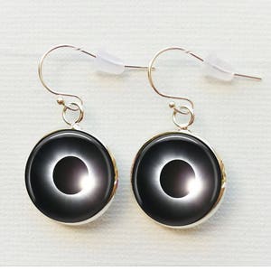 Puede incluir: Un par de pendientes de plata con colgantes circulares negros que presentan una imagen blanca de un eclipse solar.