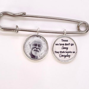 May include: A silver safety pin with two round charms. One charm has a black and white photo of a man with a beard. The other charm has the text "Those we love don't go Away They Walk beside us Everyday".