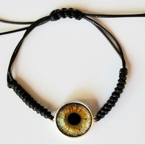 May include: A black cord bracelet featuring a central circular charm with a realistic eye design. The eye has a black pupil surrounded by a detailed iris in shades of golden yellow, brown, and green. The bracelet uses a macrame knotting technique.