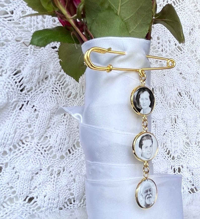 May include: A gold safety pin with a chain attached to three round photo charms. The photos are black and white and feature three different people.
