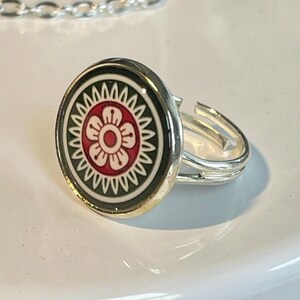 May include: A silver-toned adjustable ring featuring a circular centerpiece with a dark green border, a white serrated ring, and a central red flower motif with white petals. The metal band is split into two thin parallel wires at the sides.