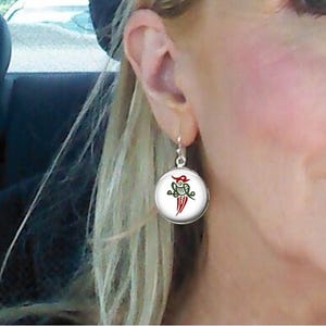May include: Round silver earrings with a white background and a red and green design. The earrings are hanging from a person's ear.