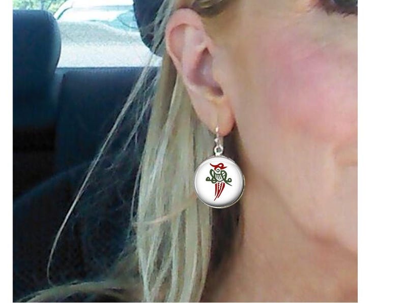 May include: Round silver earrings with a white background and a red and green design. The earrings are hanging from a person's ear.