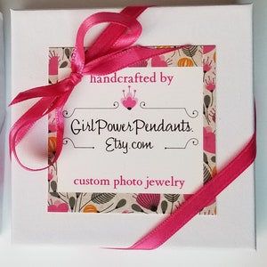 May include: A square white gift box tied with a bright pink satin ribbon. A central label features a floral pattern border and the text: handcrafted by Girl Power Pendants. Etsy.com custom photo jewelry, with a small pink heart icon above the text.