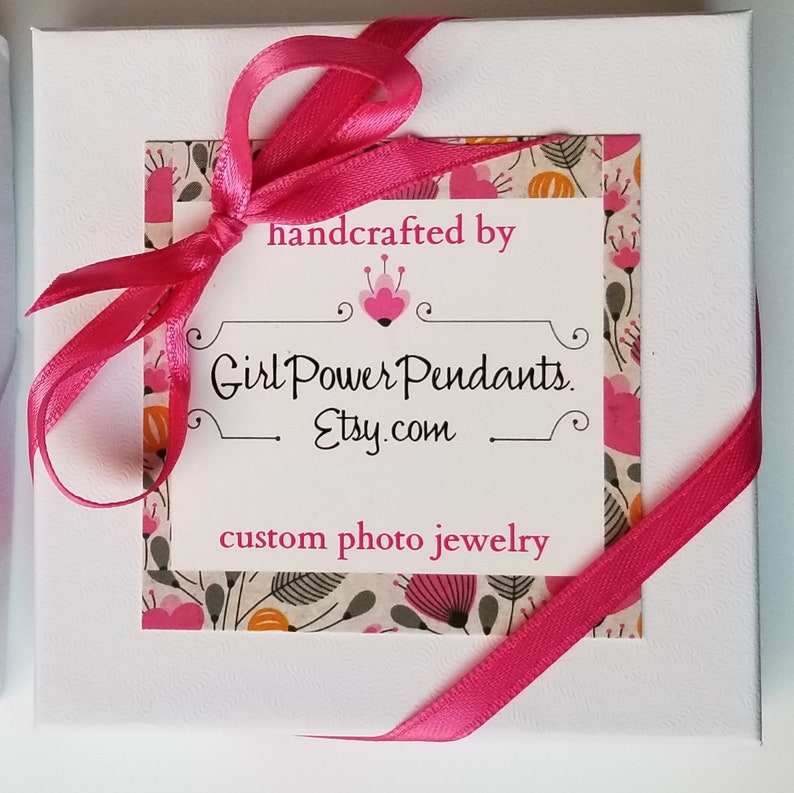 May include: A square white gift box tied with a bright pink satin ribbon. A central label features a floral pattern border and the text: handcrafted by Girl Power Pendants. Etsy.com custom photo jewelry, with a small pink heart icon above the text.