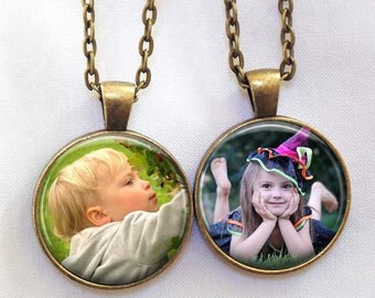 May include: Two bronze necklaces with round glass pendants featuring photos of children. The pendant on the left shows a young boy with blonde hair looking to the right. The pendant on the right shows a young girl wearing a witch costume with a pink and black hat, looking at the camera.