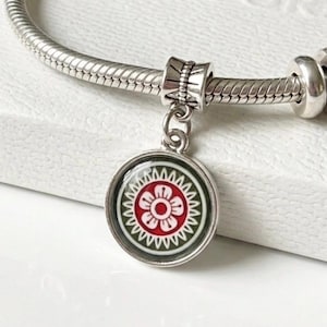 Puede incluir: Una pulsera de plata con un colgante redondo. El colgante presenta un diseño floral rojo y blanco sobre un fondo verde. La pulsera tiene un diseño de cadena de serpiente y está adornada con cuentas decorativas adicionales.