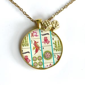 May include: A gold-toned circular pendant necklace featuring a collage of colorful Mahjong tile illustrations, including a red bird, green bamboo, and Chinese characters. Two small gold-toned dice charms are attached to the top of the pendant.