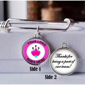 Peut inclure: Un bracelet extensible en argent avec un charme composé de deux disques ronds bordés d'argent. Le premier disque a un fond rose avec un motif de fleur stylisé et le texte "YOUR LOGO GOES HERE". Le deuxième disque a un fond blanc avec le texte "Thanks for being a part of our team!" en noir.