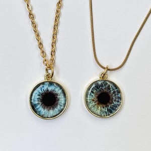 May include: Two gold-toned necklaces with round pendants featuring detailed eye designs. One eye is blue, the other is green and blue, both with black pupils. The pendants are suspended from delicate gold chains.
