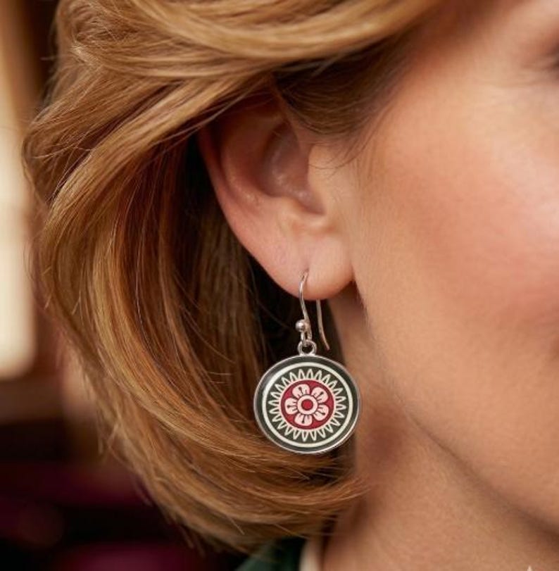 May include: Silver drop earrings with a circular design. Each earring displays a red and white floral pattern on a black background, framed by a white border. The earrings are suspended from a simple hook.