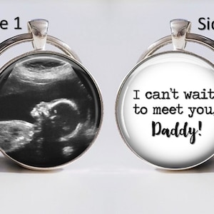 May include: A silver keychain with two sides. One side features a black and white ultrasound image of a baby. The other side has white text on a black background that reads "I can't wait to meet you, Daddy!"