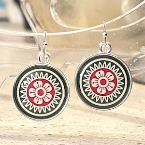 May include: A pair of circular drop earrings. Each earring displays a red and white floral pattern on a black base, framed by a silver-toned metal border. The earrings are suspended from simple hook fastenings.