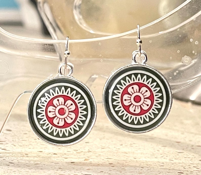 May include: A pair of circular drop earrings. Each earring displays a red and white floral pattern on a black base, framed by a silver-toned metal border. The earrings are suspended from simple hook fastenings.