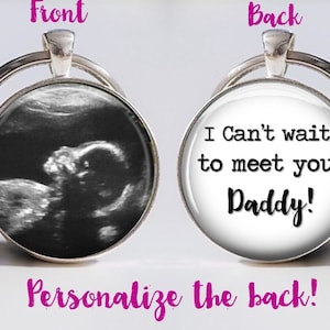 May include: A silver keychain with a round glass charm. The front of the charm has a black and white ultrasound image of a baby. The back of the charm has the text "I Can't wait to meet you, Daddy!" in black text on a white background.