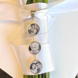 May include: A silver-toned metal pendant featuring three circular frames, each containing a black and white photograph. The pendant is attached to a white satin ribbon wrapped around a bundle of green stems, creating a decorative memorial charm.