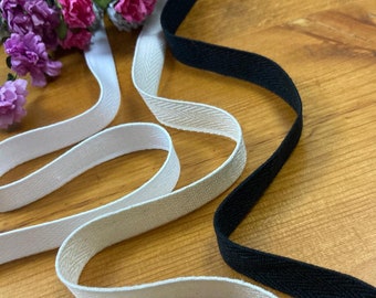May include: Two rolls of white and black cotton twill tape. The tape is woven with a herringbone pattern. The tape is on a wooden surface.