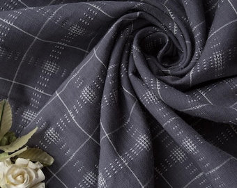 May include: Close-up of a dark gray fabric with a white grid pattern. The fabric is draped and folded, creating a textured appearance. A white rose and green leaves are in the lower left corner. The fabric is likely used for clothing or home decor.
