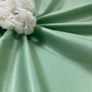 May include: A smooth, light green fabric with a subtle sheen. The fabric is draped and folded, creating soft curves and folds. A cluster of white silk flowers is placed on the fabric.