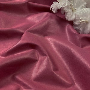 May include: A close-up of a smooth, pink fabric with a subtle sheen. The fabric is draped and folded, creating soft curves and folds. A white flower is partially visible in the top right corner of the image.