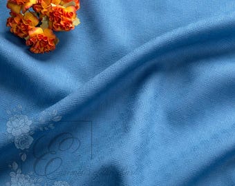 May include: A close-up of a light blue fabric with a subtle woven pattern. A small cluster of orange and yellow flowers sits in the upper left corner. The fabric is draped, creating soft folds. A floral design is in the lower left corner.