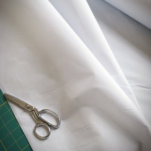 Puede incluir: Un rollo de tela blanca con un par de tijeras plateadas sobre una estera de corte verde. La tela está doblada por la mitad y las tijeras están abiertas.