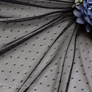 May include: Close-up of black dotted tulle fabric. The delicate netting features small, evenly spaced dots. The fabric is draped, creating folds and shadows. A cluster of blue flowers is visible in the upper right corner.