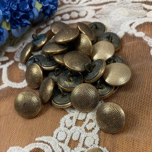 May include: A pile of 20 gold-toned metal buttons with a textured surface. The buttons are round and have a raised center. They are arranged in a pile on a white lace fabric.