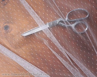 Può includere: Tessuto di tulle bianco a pois con bordo a nastro, tagliato con forbici argentate. Il tessuto è appoggiato su una superficie di legno. L'indirizzo del sito web "www.EnsemblesOfThePast.com" è visibile in basso nell'immagine.