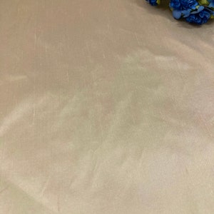 May include: A close-up of a smooth, light beige fabric with a subtle sheen. The fabric has a slightly wrinkled texture. The fabric is displayed on a flat surface.