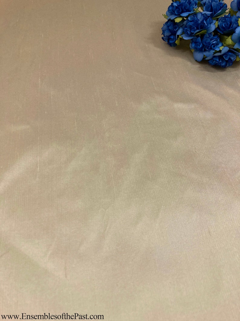 May include: A close-up of a smooth, light beige fabric with a subtle sheen. The fabric has a slightly wrinkled texture. The fabric is displayed on a flat surface.