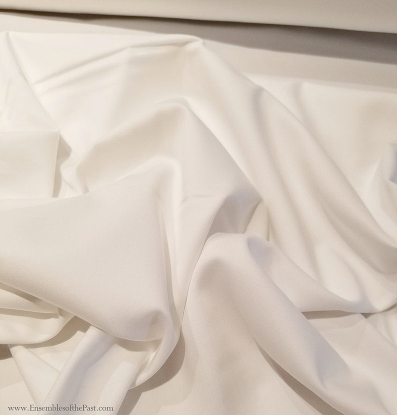 May include: White fabric with a smooth texture. The fabric is folded and draped, showing its soft and flowing nature.