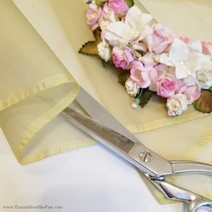 Può includere: Un paio di forbici d'argento con un tessuto trasparente giallo pallido con bordo dorato. Il tessuto è decorato con un gruppo di fiori rosa e bianchi.