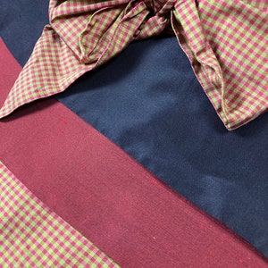 Puede incluir: Primer plano de un lazo de vichy rojo y verde sobre un fondo de tela azul oscuro y burdeos. La tela est&aacute; doblada en diagonal. El texto "www.EnsemblesofthePast.com" es visible en la esquina inferior derecha de la imagen.