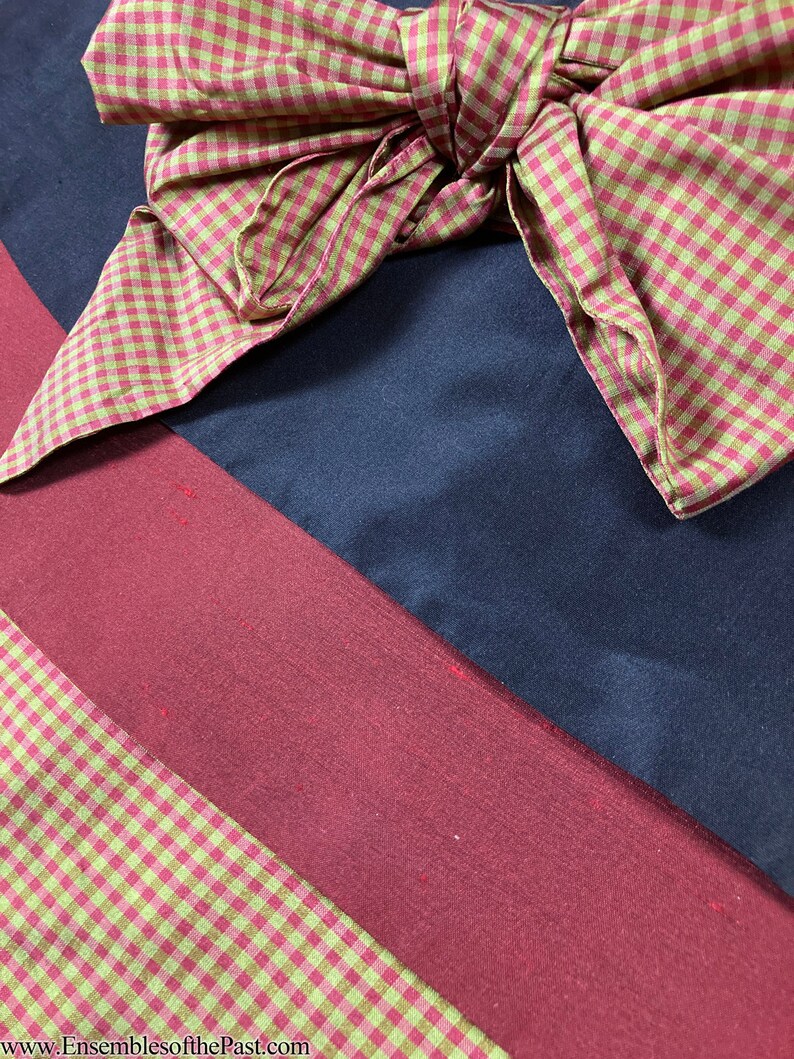 Puede incluir: Primer plano de un lazo de vichy rojo y verde sobre un fondo de tela azul oscuro y burdeos. La tela est&aacute; doblada en diagonal. El texto "www.EnsemblesofthePast.com" es visible en la esquina inferior derecha de la imagen.