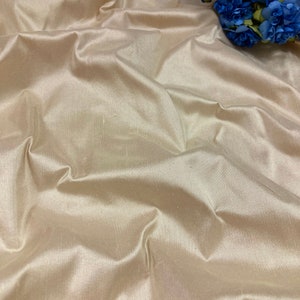 May include: A close-up of a smooth, ivory-coloured fabric with a subtle sheen. The fabric is draped and folded, creating soft, flowing lines. The fabric is likely silk or satin.