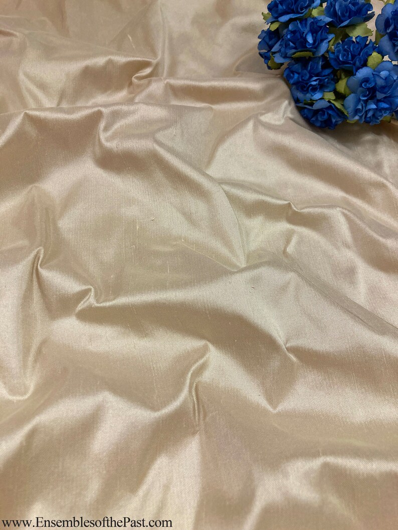 May include: A close-up of a smooth, ivory-coloured fabric with a subtle sheen. The fabric is draped and folded, creating soft, flowing lines. The fabric is likely silk or satin.