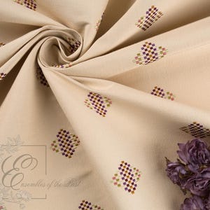 May include: Cream-colored fabric with a repeating pattern of small, embroidered geometric shapes in purple, green, and brown. The fabric is draped, showing folds and texture. A cluster of dried purple flowers is in the lower right corner. The text "Ensembles of the Past" is in the lower left corner.