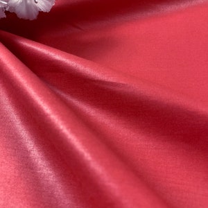 Puede incluir: Primer plano de una tela roja lisa y sedosa con un ligero brillo. La tela est&aacute; drapeada y doblada, creando curvas y pliegues suaves. Una flor blanca es visible en la esquina superior izquierda de la imagen.