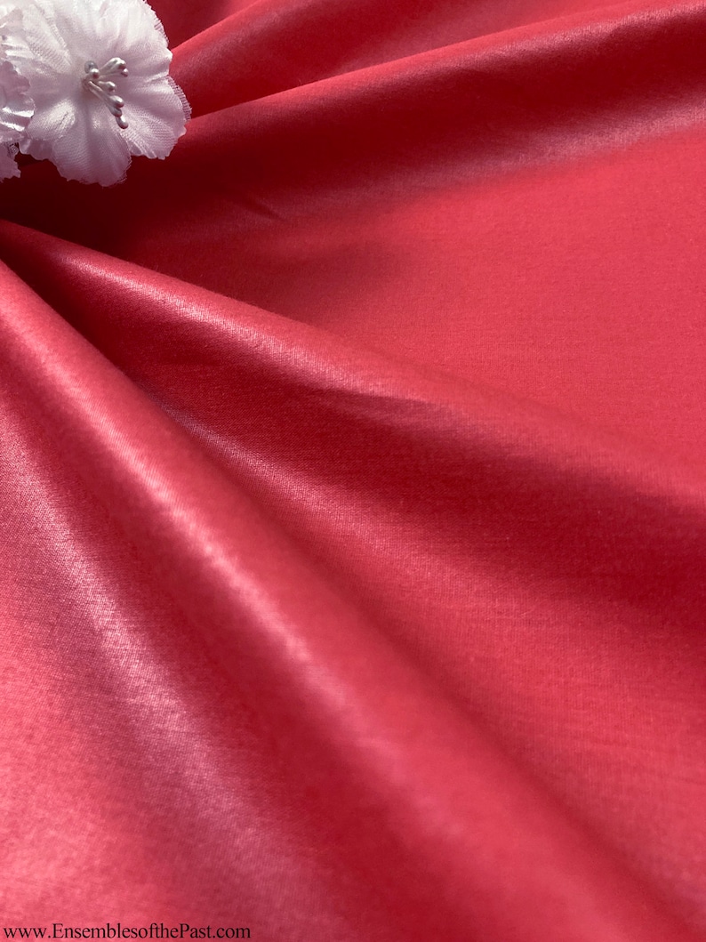 Puede incluir: Primer plano de una tela roja lisa y sedosa con un ligero brillo. La tela est&aacute; drapeada y doblada, creando curvas y pliegues suaves. Una flor blanca es visible en la esquina superior izquierda de la imagen.