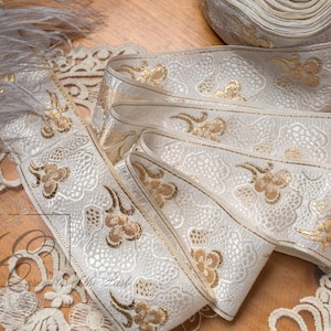 Op de afbeelding: Een rol wit lint met een metallic goudkleurig bloemmotief en een textuur die op kant lijkt. Het lint heeft afgewerkte randen en wordt getoond naast een stuk wit kanten randje en een grijze veer op een houten ondergrond.