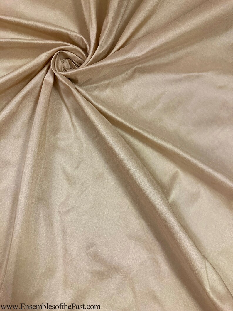 May include: A close-up of a smooth, beige fabric with a silky texture. The fabric is draped and folded, creating soft curves and folds.