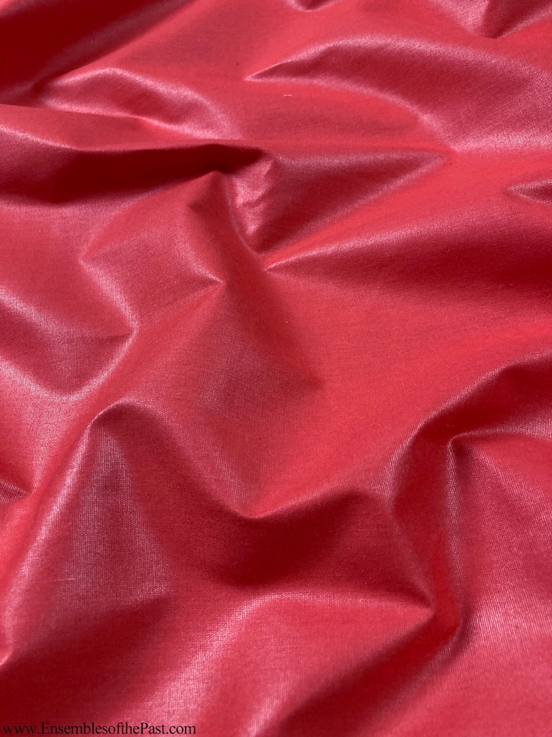 Puede incluir: Primer plano de una tela lisa de color rojo con un brillo sutil. La tela est&aacute; doblada y drapeada, creando l&iacute;neas suaves y fluidas.