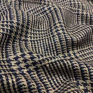 May include: Close-up of a textured fabric with a woven pattern in navy blue, light blue, and off-white. The fabric has a houndstooth design, with the weave creating a subtle, dimensional effect. The fabric is draped, showing folds and shadows.