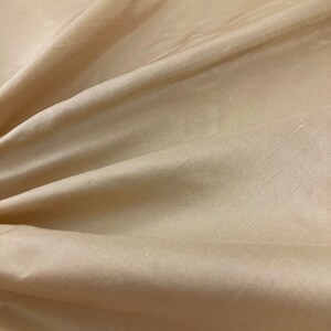 May include: A close-up of a smooth, beige fabric with a subtle sheen. The fabric is draped and folded, showcasing its texture and drape.