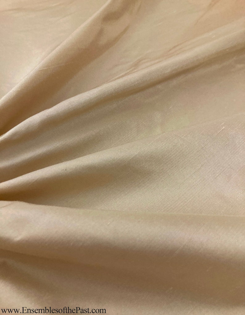 May include: A close-up of a smooth, beige fabric with a subtle sheen. The fabric is draped and folded, showcasing its texture and drape.