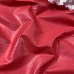 Puede incluir: Primer plano de una tela lisa de color rojo con un brillo sutil. La tela est&aacute; drapeada y doblada, creando l&iacute;neas suaves y fluidas. Una flor blanca se coloca en la parte superior de la tela.