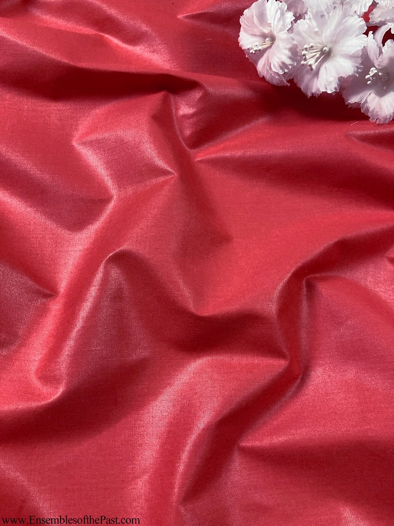 Puede incluir: Primer plano de una tela lisa de color rojo con un brillo sutil. La tela est&aacute; drapeada y doblada, creando l&iacute;neas suaves y fluidas. Una flor blanca se coloca en la parte superior de la tela.