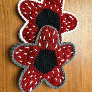 May include: Two handmade crochet Demogorgon flower coasters. The coasters are red with white accents and a black center. The top coaster has a white border, while the bottom coaster has a gray border. The coasters are placed on a wooden surface.