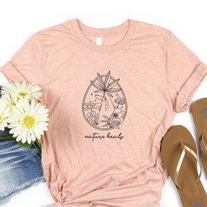 May include: A light pink t-shirt with a black line drawing of a hand holding a marijuana leaf surrounded by flowers. The text "nature heals" is printed below the drawing.